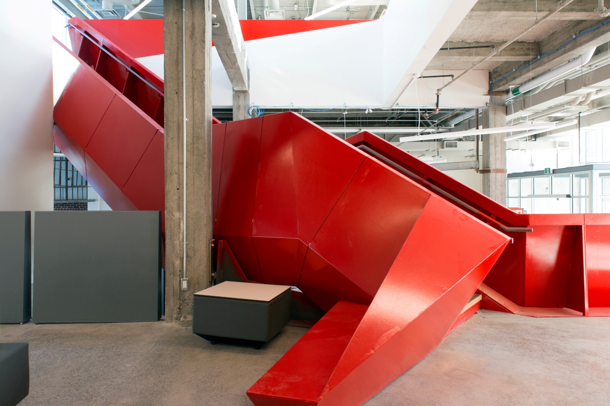 Interior photo of an imposing red metal staircase nestled in the middle of a shared community office space in what was once a former garage.